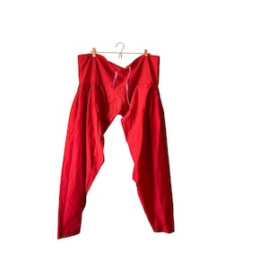 May include: Bright red loose-fitting trousers featuring a gathered waistband with a drawstring closure. The pants have a wide-leg design with pleats at the top, hanging from a wooden hanger. The fabric appears lightweight and solid in color.