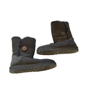 May include: A pair of black UGG boots. The boots have a button closure on the side and a fur trim at the top. The soles are black with a textured pattern. The boots are made of suede.