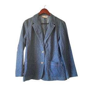 Op de afbeelding: Een spijkerblazer in een medium blauwe wassing, met een klassieke revers, twee voorzakken en een sluiting met één knoop. De jas hangt aan een houten hanger. Het label luidt "ecoluy".