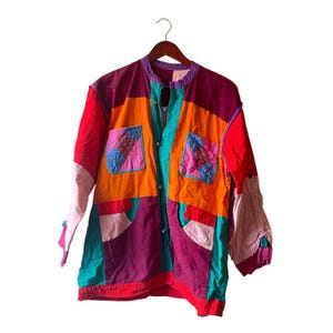 Può includere: Giacca patchwork colorata con collo tondo e maniche lunghe. La giacca presenta un mix di pannelli rossi, arancioni, viola, verde acqua e rosa. Due tasche quadrate con dettagli trapuntati sono sul davanti. La giacca è appesa a una gruccia di legno.