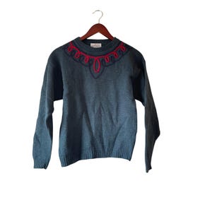 Puede incluir: Un suéter de color verde azulado con cuello redondo y mangas largas, colgado de una percha de madera. El cuello presenta un diseño decorativo en azul marino y rojo. El suéter está hecho de un material de punto suave.