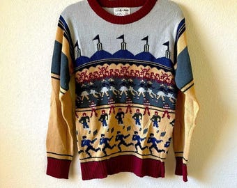 May include: A vintage crew neck sweater with a colorful pattern. The sweater features a gray upper section, a burgundy collar and cuffs, and a design of running figures and flags. The sleeves are tan and blue.