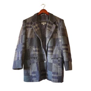 May include: A patchwork blazer in shades of blue, gray, and green hangs on a wooden hanger. The blazer features a variety of plaid and patterned fabric panels. It has a notched collar, two front pockets, and a single-button closure.
