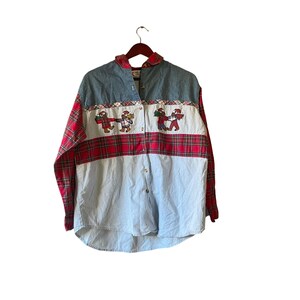 Könnte beinhalten: Ein langärmliges Button-Down-Hemd mit Jeanspasse und Ärmeln, rot karierten Ärmeln und einem weiß-rot karierten Band über der Brust. Das Hemd hat gestickte Figuren mit Weihnachtsmützen. Das Hemd hängt an einem Holzbügel.