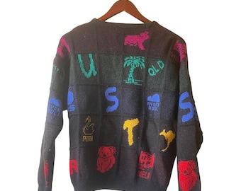 May include: Dark grey sweater with colourful embroidered designs. Features a patchwork design with letters, a palm tree, a koala, and other Australian-themed images. The sweater is hanging on a wooden hanger.