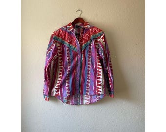 May include: A button-down shirt with a colorful geometric pattern. The shirt features a mix of red, purple, blue, and white vertical stripes and triangular accents. It has a collared neckline and long sleeves, hanging on a wooden hanger.