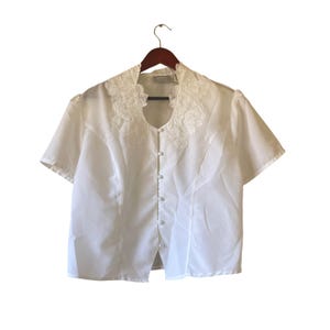 May include: A white short-sleeved blouse with a decorative floral lace collar and button-down front. The blouse is hanging on a wooden hanger against a white background. The fabric appears to be lightweight and semi-sheer.
