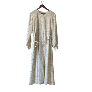 May include: A long, button-down dress with a floral pattern in shades of lavender and green on a cream background. The dress has long sleeves, a matching belt, and is hanging on a wooden hanger.