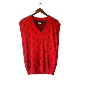 Peut inclure: Un gilet de pull rouge à col en V avec un motif de carrés noirs. Le gilet est suspendu à un cintre en bois sur un fond blanc. Le gilet de pull est un vêtement sans manches.