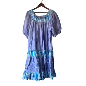 May include: A lilac-colored dress with short, puffed sleeves and a square neckline. The dress features a tiered skirt with blue and white patterned accents. The top of the dress has a floral design. The dress is hanging on a wooden hanger.