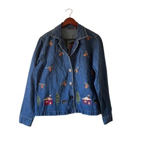 Könnte beinhalten: Blaue Jeansjacke mit gestickten Herbstblättern, Hütten und Bären. Die Jacke hat einen Kragen, eine Knopfleiste und lange Ärmel. Die Jacke hängt an einem Holzkleiderbügel.