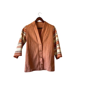 May include: A vintage peach cardigan with a V-neck and button fastening. The sleeves have a striped pattern in cream, orange, and brown. The cardigan is displayed on a wooden hanger against a white backdrop.