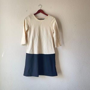May include: A cream-colored dress with a navy blue skirt, hanging on a wooden hanger. The dress has three-quarter sleeves and a round neckline. The skirt portion is a solid, dark blue color. The dress appears to be made of a soft, comfortable material.