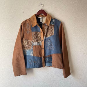 May include: A brown and blue suede jacket with a patchwork design. The jacket features embroidered floral and swirl patterns. It has a button-up front and a collared neckline. The sleeves are long and brown.