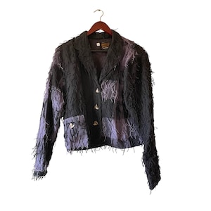 May include: A black blazer with a textured, fringed design and patches of lavender fabric. The jacket has three decorative buttons and two pockets. It is hanging on a wooden hanger.
