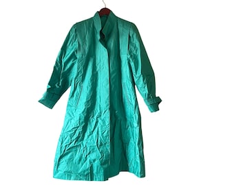 May include: A long, teal-colored trench coat hanging on a wooden hanger. The coat features a high collar, pleated shoulder details, and a button closure. The sleeves have buttoned cuffs. The coat is made of a shiny, lightweight material.