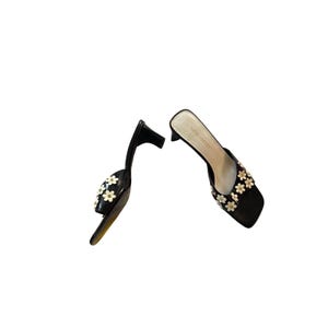 May include: Black Nine West heeled sandals with white daisy accents. The open-toe mules have a small heel and a square toe. The sandals are decorated with white daisy-shaped floral embellishments.
