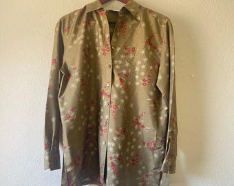 Puede incluir: Camisa de manga larga verde oliva con un delicado estampado floral en blanco y rosa. La camisa está abotonada y colgada de una percha de madera sobre un fondo neutro.