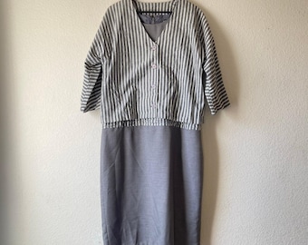 Puede incluir: Conjunto de dos piezas. La parte superior es una blusa abotonada de manga corta con rayas verticales grises y blancas. La parte inferior es una falda lápiz gris lisa hasta la rodilla. El vestido está colgado en una percha de madera.