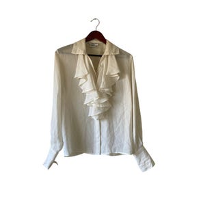 May include: Cream-coloured long-sleeved blouse with a ruffled front and buttoned cuffs. The blouse is hanging on a wooden hanger against a white background. The collar is a classic point collar.