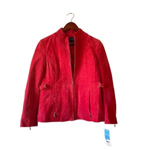 May include: A red suede jacket with a stand-up collar, long sleeves, and a front zipper. The jacket has two small pockets and decorative buckles on the sides. It is hanging on a wooden hanger.