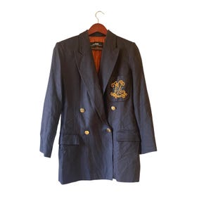 May include: Navy blue double-breasted blazer with gold-colored buttons and an embroidered emblem on the pocket. The blazer has a classic design with a notched lapel and a tailored fit. The interior lining is a warm orange color.