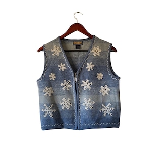 May include: A blue and gray sleeveless vest with a snowflake pattern. The vest has a zipper closure and is hanging on a wooden hanger. The snowflakes are white and embroidered on the front of the vest.