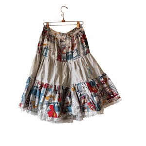 Könnte beinhalten: Ein mehrlagiger Vintage-Rock mit einem skurrilen Druck mit roten, blauen und weißen Illustrationen auf hellgrauem Hintergrund. Der Rock hat eine weiße Spitzenborte am unteren Saum und hängt an einem Holzkleiderbügel.