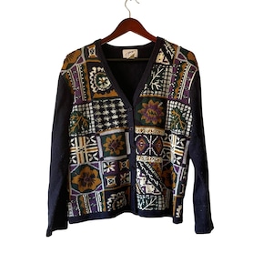 May include: A vintage cardigan sweater with a patchwork design. The sweater features a V-neck, button-up front, and long sleeves. The patchwork design includes floral, geometric, and leaf patterns in shades of navy, purple, green, and gold.