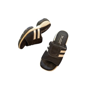 Op de afbeelding: Een paar zwarte slippers met twee witte strepen over de bovenste band. De slippers hebben een dikke, beige zool en de merknaam Mudd is in het wit gedrukt op het zwarte voetbed van de slipper die aan de rechterkant is geplaatst.
