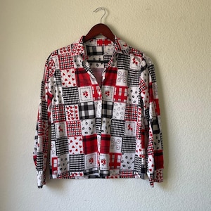 Puede incluir: Una camisa de manga larga con un diseño de patchwork. La camisa presenta cuadrados de patrones rojos, negros y blancos, incluyendo corazones, cuadros y motivos florales. La camisa está colgada en una percha de madera contra una pared neutra.