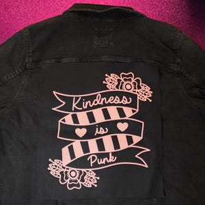 May include: A black denim jacket featuring a large rectangular patch on the back. The patch displays a light pink graphic of a ribbon with black stripes, two hearts, and floral accents. The text "Kindness is Punk" is printed in a script font.
