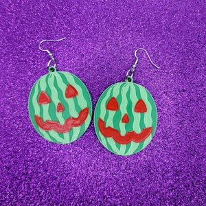 May include: A pair of green watermelon-shaped earrings with a jack-o'-lantern face design. The earrings are made of plastic and have silver hooks.