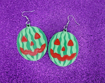 May include: A pair of green watermelon-shaped earrings with a jack-o'-lantern face design. The earrings are made of plastic and have silver hooks.
