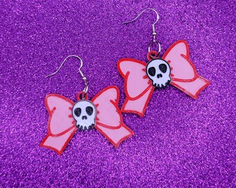May include: A pair of pink bow earrings with a white skull charm. The earrings are made of plastic and have silver hooks.