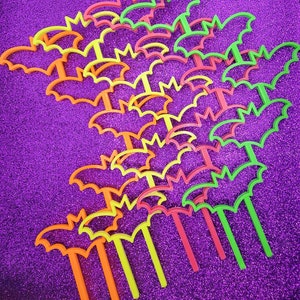 May include: A collection of colorful plastic bat-shaped decorations on sticks. The bats are in various shades of orange, yellow, and green, and are arranged in a random pattern on a purple sparkly background.