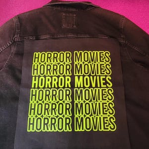 May include: Black denim jacket with a large black patch on the back. The patch features the words "HORROR MOVIES" in a repeating pattern of bright neon green block letters. The jacket has a classic collar and button closures.