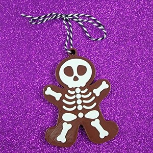 May include: A brown gingerbread man ornament with a white skeleton design. The ornament has a white string attached to the top.
