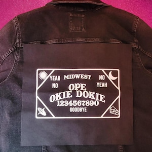 May include: Black denim jacket with a large, black rectangular patch on the back. The patch features white text and graphics, including the words "Yeah Midwest," "Ope Okie Dokie," and numbers 1-0.