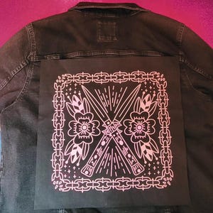 May include: Black denim jacket with a large, square patch on the back. The patch features a pink and white design of crossed knives, flowers, and chain-link borders. The jacket is a dark wash.