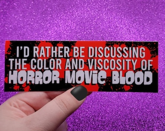 Puede incluir: Una pegatina rectangular con el texto "I'D RATHER BE DISCUSSING THE COLOR AND VISCOSITY OF HORROR MOVIE BLOOD" en letras blancas sobre un fondo negro y rojo. La pegatina está sujeta por una mano con una uña pintada de negro.