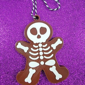 May include: Brown gingerbread cookie ornament shaped like a skeleton with a white skull and white bones. The ornament is hanging from a black and white string.