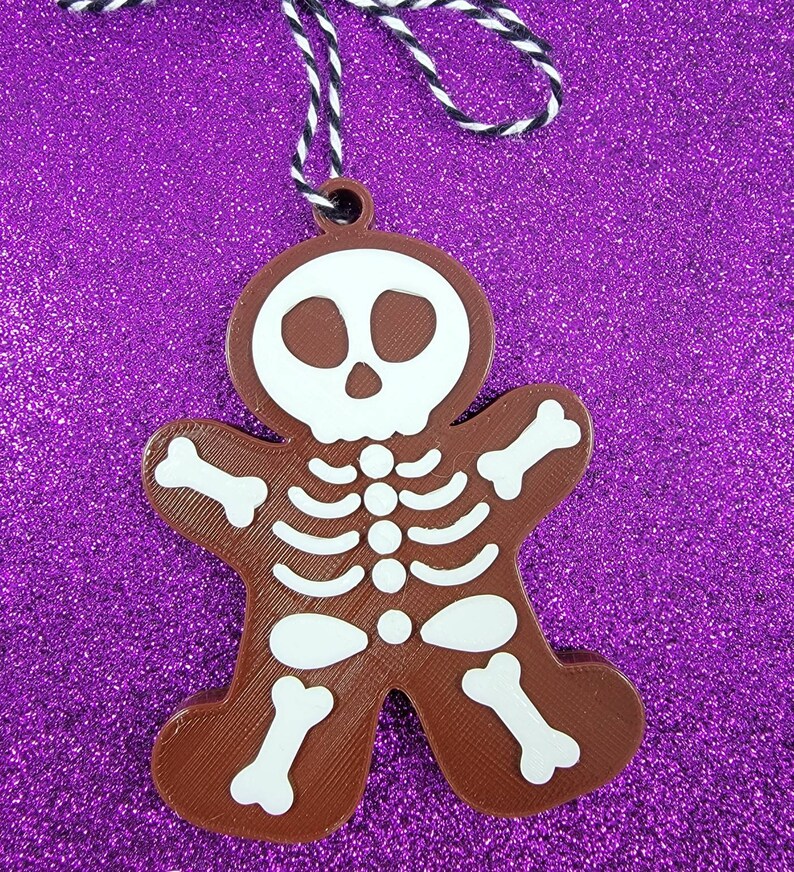 May include: Brown gingerbread cookie ornament shaped like a skeleton with a white skull and white bones. The ornament is hanging from a black and white string.
