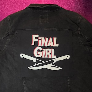 May include: A black denim jacket featuring a large rectangular patch on the back. The patch is black with the white text "FINAL GIRL" in a bold, stylized font with red drop shadows. Below the text are two white crossed kitchen knives.