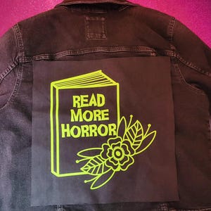 May include: Black denim jacket with a large black square patch on the back. The patch features a neon green illustration of an open book with the words "READ MORE HORROR" and floral accents.
