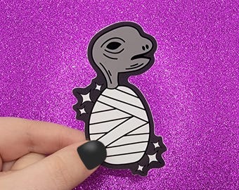 Peut inclure: Un autocollant représentant une tortue de dessin animé enveloppée de bandages. La tortue a une tête et un cou gris, avec des bandages blancs et des contours noirs. L'autocollant est posé sur un fond violet scintillant.