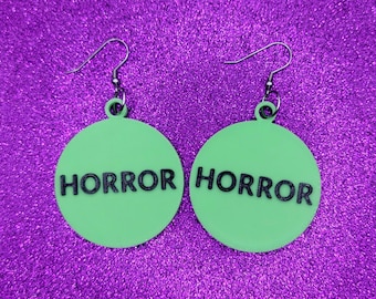 May include: A pair of green round earrings with the word "HORROR" printed in black on each earring.