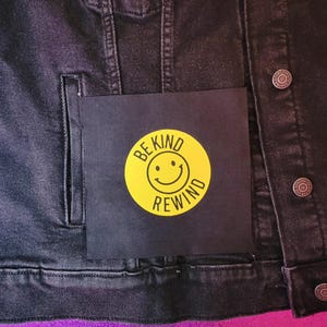 May include: Black denim jacket with a square patch featuring a yellow circle with a smiling face and the words "BE KIND REWIND" in black. The jacket has silver buttons and a pocket.