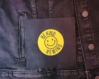 May include: Black denim jacket with a square patch featuring a yellow circle with a smiling face and the words "BE KIND REWIND" in black. The jacket has silver buttons and a pocket.