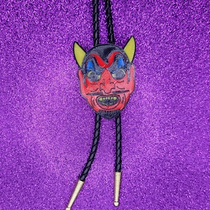 May include: A red devil face bolo tie with black braided cords and gold-colored tips. The devil face has yellow horns, black eyes, and a white mustache.